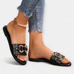 Black slide sandals with round toe and wide gold-buckled straps, low flat sole