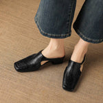 Pair of black leather mules with square toe and low heels on a beige fabric surface.