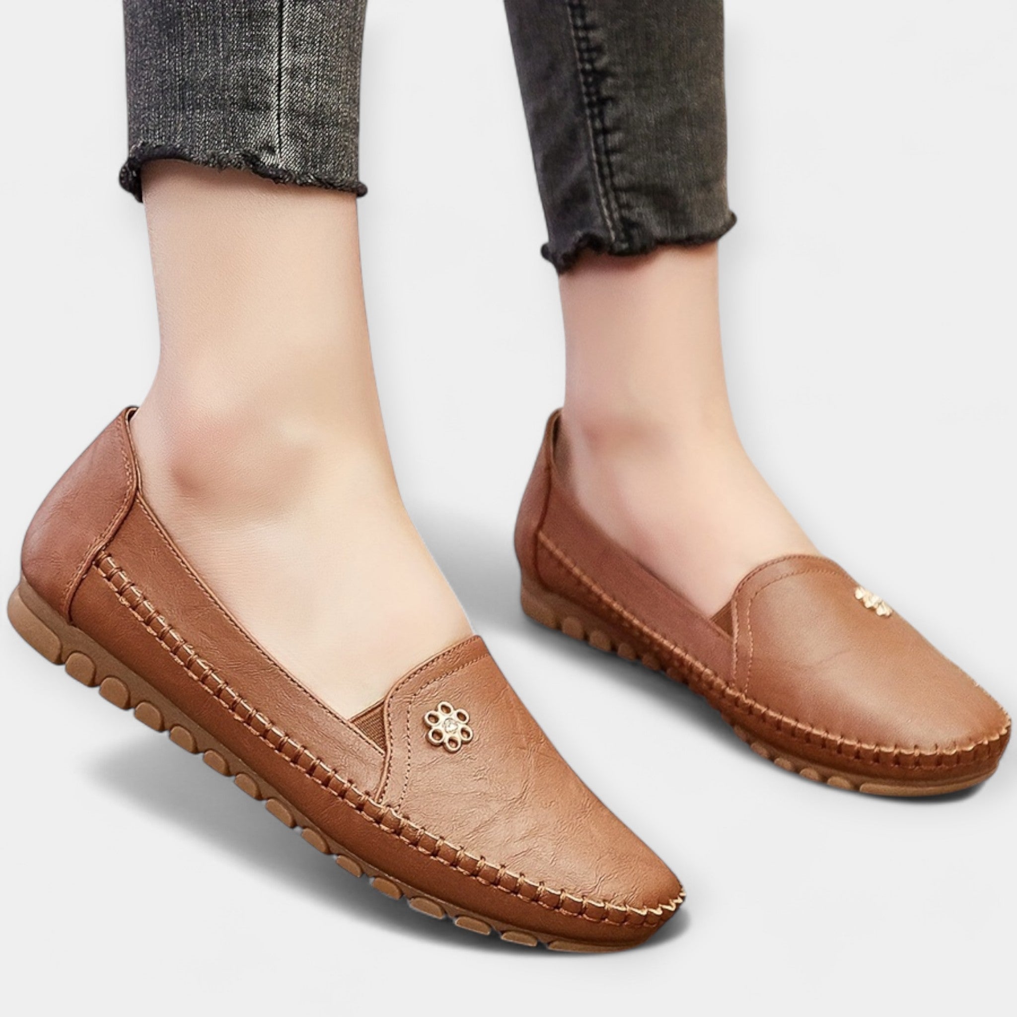Brown slip-on loafers with soft sole, stitched edges, and small floral emblem on upper