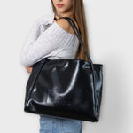 Black leather-look tote bag held by model, wide top opening and stitched seams